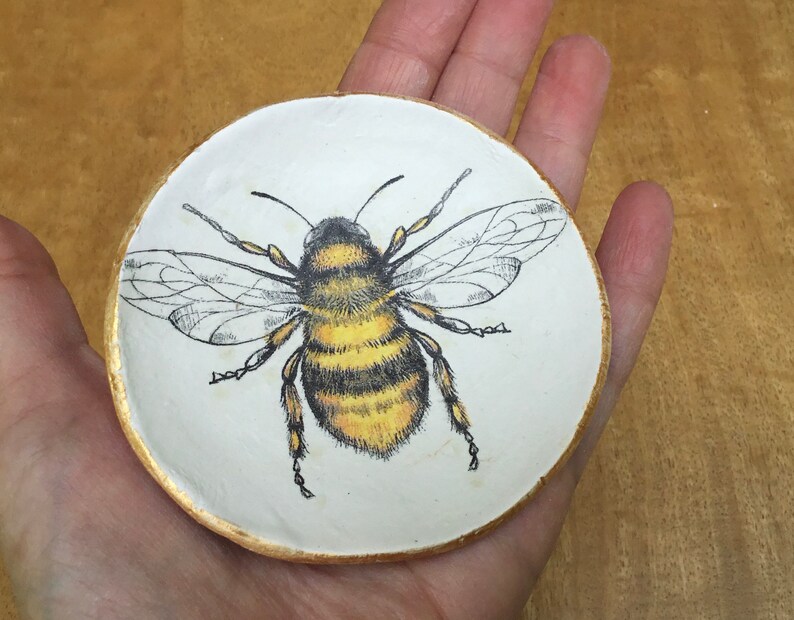 Op de afbeelding: Een wit keramiek sieradenbakje met een gouden rand heeft een handgeschilderde illustratie van een hommel met zwarte en gele strepen.