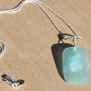 May include: A silver chain necklace with a light blue frosted glass pendant. The pendant is rectangular in shape and has a slightly rounded edge.