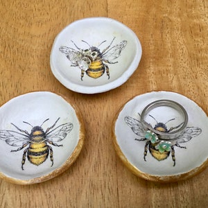 May include: Three white ceramic ring dishes with a gold rim and a bee design. Each dish features a different bee with a black and yellow striped body and brown wings. One dish has a ring with a silver band and two green flowers on it.