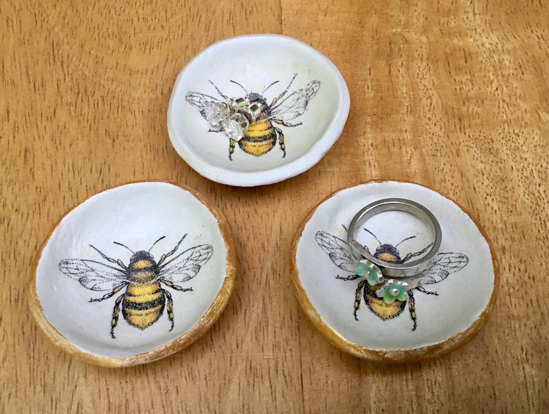 May include: Three white ceramic ring dishes with a gold rim and a bee design. Each dish features a different bee with a black and yellow striped body and brown wings. One dish has a ring with a silver band and two green flowers on it.