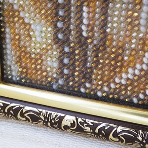 Pu&ograve; includere: Primo piano di un'opera d'arte con perline incorniciata con una cornice dorata e una bordura in legno marrone. Le perline sono in tonalit&agrave; dorate, marroni e bianche, creando uno schema testurizzato.