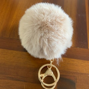 May include: A fluffy beige pom pom keychain with a gold metal key ring.