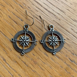 Puede incluir: Un par de pendientes de plata con forma de brújula con un diseño detallado. La brújula presenta las direcciones cardinales N, S, E y W.