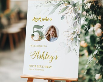 Può includere: Un cartello di compleanno rettangolare bianco su un cavalletto di legno con foglie ad acquerello verdi e dettagli dorati. Il testo recita: Look who's 50, WELCOME TO Ashley's 50TH BIRTHDAY, DEC 10 - NEW YORK. Una foto circolare è integrata nel numero 50.