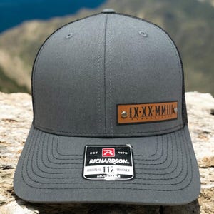May include: A grey and black trucker hat with a brown leather patch that says "IX-XX-MMIII RYANNARACER". The hat has a black mesh back and an adjustable strap with a black buckle. The hat also has a small white label that says "EST. 1970 RICHARDSON. ORIGINAL 117 TRUCKER ADJUSTABLE".