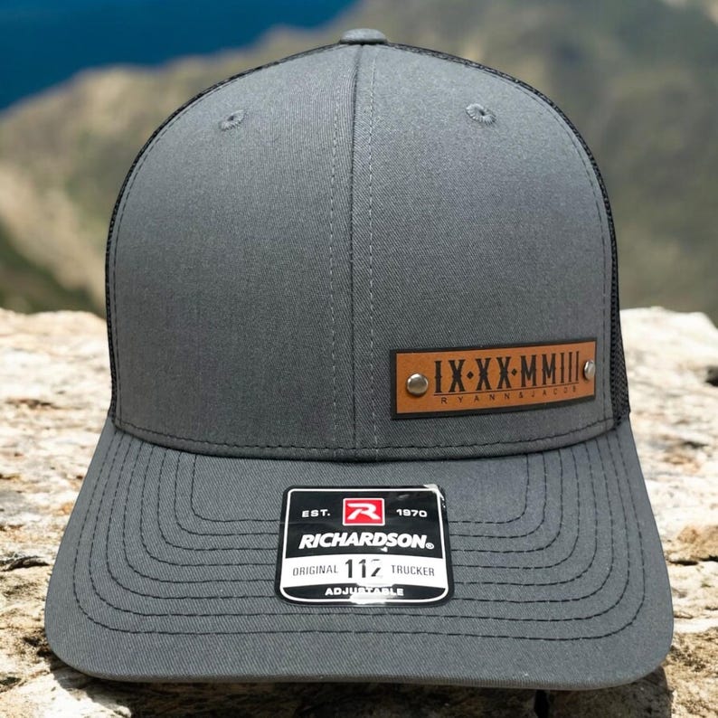 May include: A grey and black trucker hat with a brown leather patch that says "IX-XX-MMIII RYANNARACER". The hat has a black mesh back and an adjustable strap with a black buckle. The hat also has a small white label that says "EST. 1970 RICHARDSON. ORIGINAL 117 TRUCKER ADJUSTABLE".