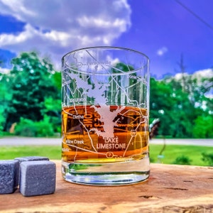 May include: A clear glass with a brown liquid inside. The glass has an etched design of Lake Limestone, a map of the lake, and surrounding towns. The glass is sitting on a wooden surface with gray stone coasters.
