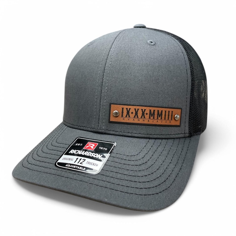 May include: A grey and black trucker hat with a brown leather patch that reads "IX-XX-MMIII RYAN & JACOBS". The hat is adjustable and has a Richardson 112 label.