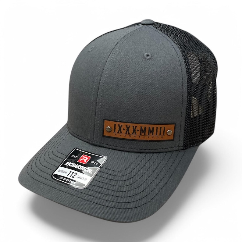 May include: Gray and black trucker hat with a brown leather patch that says "IX-XX-MMIII" in Roman numerals. The hat is adjustable and has a mesh back.