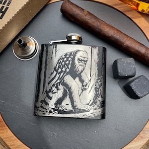 May include: A stainless steel flask with a Bigfoot graphic wearing an American flag. The flask has a silver cap and a small funnel. A cigar and two square whiskey stones are also present.