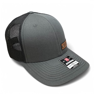 May include: A grey and black trucker hat with a brown leather patch that says "AXE" on it. The hat has a black mesh back and a grey front. The hat is made by Richardson 112.