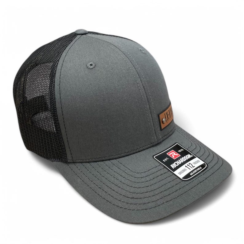 May include: A grey and black trucker hat with a brown leather patch that says "AXE" on it. The hat has a black mesh back and a grey front. The hat is made by Richardson 112.