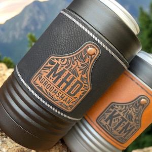 May include: Two black and brown leather can coolers with a metal top. The coolers have a decorative metal tag with a floral design and the text "MHD GROOMSMAN" and "KIM".