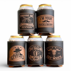 May include: A stack of six black leatherette can coolers with brown leatherette patches. Each cooler has a different design and text, including "Forgive Me If I Don't Shake Hands" and "I'm Your Huckleberry."