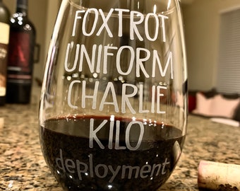 May include: A clear glass wine glass with the words "FOXTROT UNIFORM CHARLIE KILO DEPLOYMENT" etched on the side. The glass is filled with red wine.