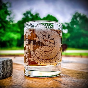 May include: A clear glass tumbler with a rattlesnake design and the words "Don't Tread On Me" etched on the side. The glass is filled with a brown liquid.