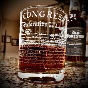 May include: A clear glass tumbler filled with amber liquid, featuring white text of the Declaration of Independence. The word "CONGRESS" is at the top. An Old Forester whiskey bottle is in the background.