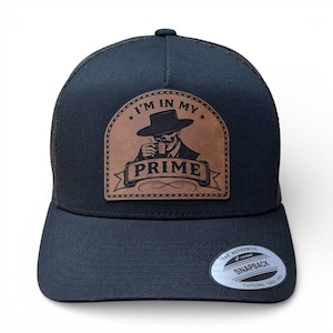 May include: Black trucker hat with a curved brim and mesh back. A brown leather patch features a skull in a suit drinking from a cup, with the text "I'M IN MY PRIME" and "PRIME". The hat has a snapback closure.