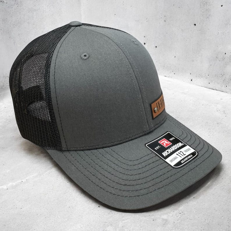 May include: A grey and black trucker hat with a brown leather patch that says "1112" on it. The hat has a black mesh back and a grey front.