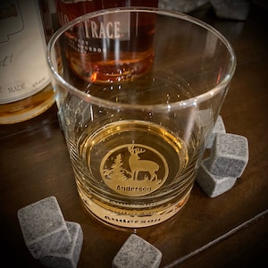 May include: A personalized whiskey glass with a gold deer silhouette and the name "Anderson" engraved on the front. The glass is filled with amber liquid and sits on a wooden surface with gray stone coasters.