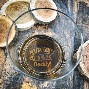 May include: A clear glass with a gold inscription that reads "You're going to be a Daddy!" The inscription is surrounded by a banner with a footprint on either side. The glass is sitting on a wooden surface.