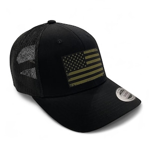 May include: Black trucker hat featuring a distressed American flag patch in olive green and tan. The hat has a mesh back and a curved brim. A small, circular tag is on the brim.
