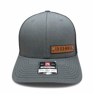 May include: A grey and black trucker hat with a brown leather patch that says "IX-XX-MMille" and "TANRACALZA". The hat is adjustable and has a Richardson logo on the back.