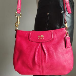 May include: A bright pink leather Coach shoulder bag with a matching strap and gold-tone hardware. The bag has a flap closure and a small tag. The Coach logo is visible on the front.