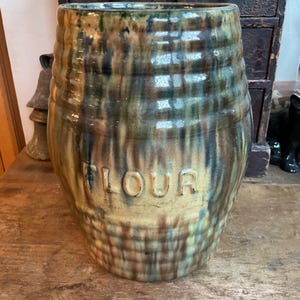 May include: A cylindrical ceramic flour crock featuring a ribbed texture and a variegated glaze in shades of green, brown, and tan. The word FLOUR is embossed in capital letters on the front, with vertical drip patterns throughout the surface.