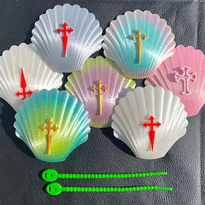 May include: A collection of scallop shell-shaped ornaments in various colors, including white, pink, and teal, each adorned with a decorative cross. Two green, flexible straps are at the bottom. The shells have a textured, ridged design.