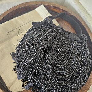 Könnte beinhalten: Eine schwarze Handtasche mit aufwendigen Perlenstickereien, facettierten schwarzen Perlen und mehreren hängenden Quasten. Die Tasche hat einen Kordelzug mit zwei runden Perlenknebeln und einen Tragegriff, dazu ein beigefarbener Stoffbeutel.