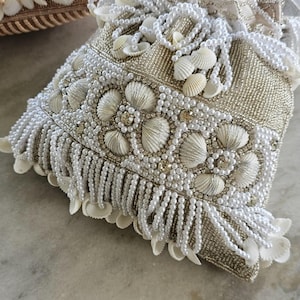 Puede incluir: Un bolso con cordón de color blanco roto adornado con conchas marinas y perlas. El bolso presenta una base tejida dorada, flecos blancos y está adornado con numerosas conchas marinas blancas y perlas de imitación. Probablemente un clutch o un bolso pequeño.