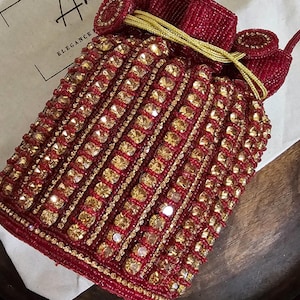 Könnte beinhalten: Rote, perlenbesetzte Handtasche mit goldenen Akzenten. Die Tasche hat einen Kordelzugverschluss und einen Griff. Die Tasche ist mit goldenen Pailletten und Perlen verziert. Die Tasche liegt auf einer Holzoberfläche.