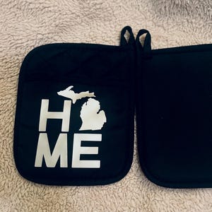 Op de afbeelding: Zwarte pannenlappen met een gewatteerd ontwerp. Eén pannenlap heeft de witte omtrek van Michigan en de woorden "HOME" en "ME" in witte letters. De andere pannenlap is effen zwart. Met ophanglus.