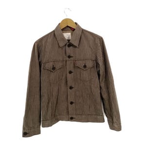 May include: Brown striped denim jacket with two chest pockets and button closures. The jacket is hanging on a wooden hanger. The jacket has a red tag on the chest pocket.