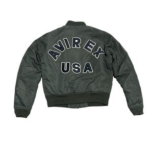 Puede incluir: Chaqueta bomber verde oscuro con la palabra "AVIREX" arqueada en la espalda en letras blancas con contorno negro, y "USA" debajo. La chaqueta tiene cuello, puños y dobladillo de canalé.