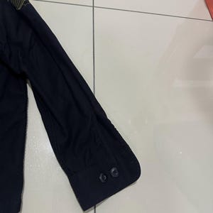 Pode incluir: Camisa preta de manga comprida com punho abotoado. A camisa apresenta listras verde-oliva no ombro e dois bot&otilde;es pretos no punho. O tecido &eacute; escuro e liso. Design de inspira&ccedil;&atilde;o militar.