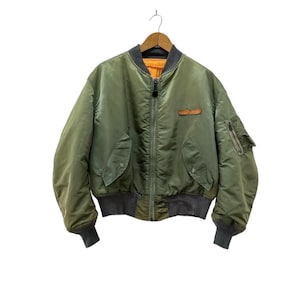 Può includere: Giacca bomber verde oliva con colletto e polsini grigi. La giacca ha una chiusura con cerniera, due tasche anteriori e una piccola tasca sulla manica sinistra. Una toppa marrone è sul petto. La giacca è appesa a una gruccia di legno.