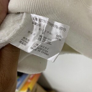 Puede incluir: Una etiqueta blanca con texto negro que detalla la composici&oacute;n, el tama&ntilde;o y el origen de la prenda. La etiqueta dice "品番 62236319(N76-609)" "品質表示" "ポリエステル 65%" "綿 35%" "サイズ" "胸囲 96~104" "身長 175~185" "発売元:" "PHONE:" "原産国: MADE IN CHINA" "(株)サイカイ" "03-3632-2101"