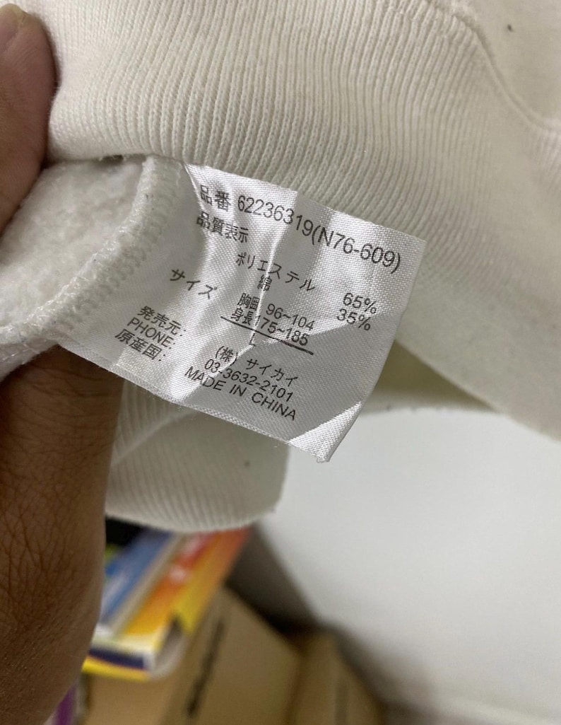 Puede incluir: Una etiqueta blanca con texto negro que detalla la composici&oacute;n, el tama&ntilde;o y el origen de la prenda. La etiqueta dice "品番 62236319(N76-609)" "品質表示" "ポリエステル 65%" "綿 35%" "サイズ" "胸囲 96~104" "身長 175~185" "発売元:" "PHONE:" "原産国: MADE IN CHINA" "(株)サイカイ" "03-3632-2101"