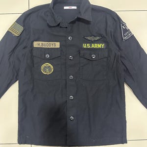 Pode incluir: Camisa preta abotoada com v&aacute;rios patches e os textos "H.BUDDYS" e "U.S. ARMY". A camisa tem gola, dois bolsos no peito e um patch da bandeira americana na manga esquerda. Feita de tecido resistente.