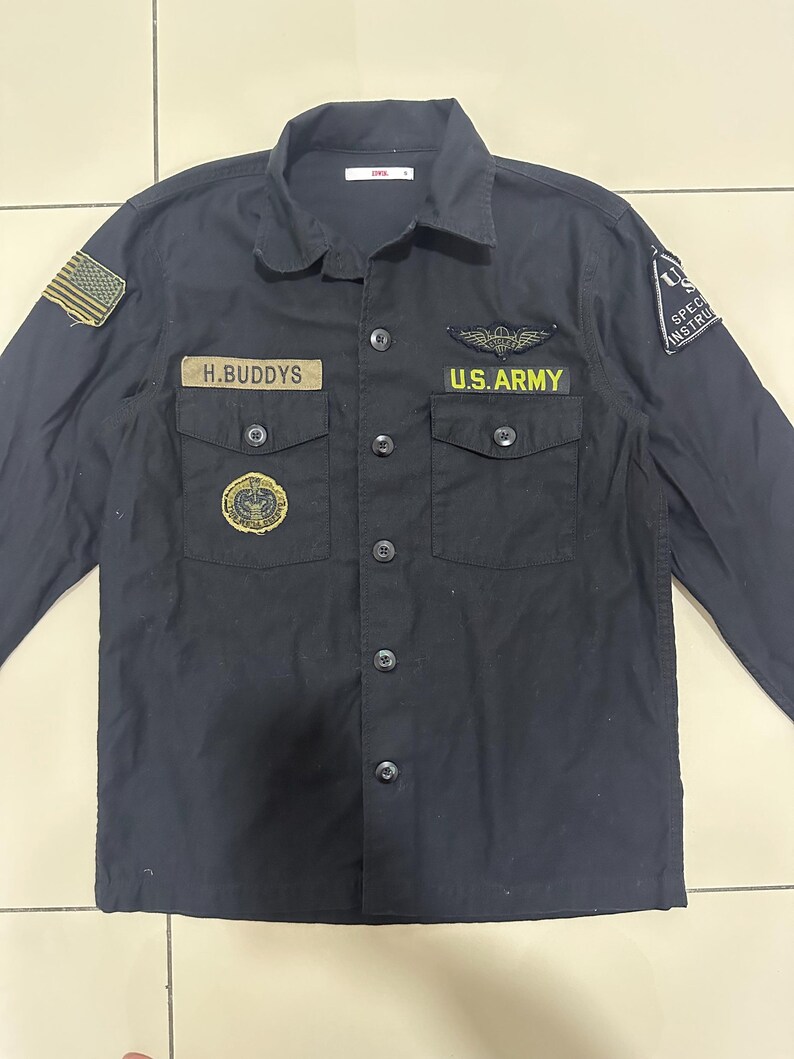 Pode incluir: Camisa preta abotoada com v&aacute;rios patches e os textos "H.BUDDYS" e "U.S. ARMY". A camisa tem gola, dois bolsos no peito e um patch da bandeira americana na manga esquerda. Feita de tecido resistente.
