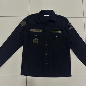 Pode incluir: Camisa preta de manga comprida com patches e ins&iacute;gnias de estilo militar. Apresenta um patch da bandeira americana, o texto "H.BUDDYS" e "U.S. ARMY" em letras douradas. A camisa tem dois bolsos no peito e uma gola.