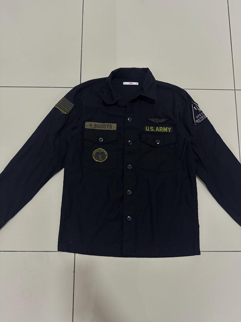 Pode incluir: Camisa preta de manga comprida com patches e ins&iacute;gnias de estilo militar. Apresenta um patch da bandeira americana, o texto "H.BUDDYS" e "U.S. ARMY" em letras douradas. A camisa tem dois bolsos no peito e uma gola.