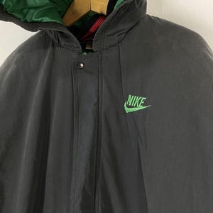 Pu&ograve; includere: Una giacca nera con cappuccio, fodera verde e un logo Nike verde sul davanti. La giacca ha una chiusura a zip e un bottone a pressione in alto.