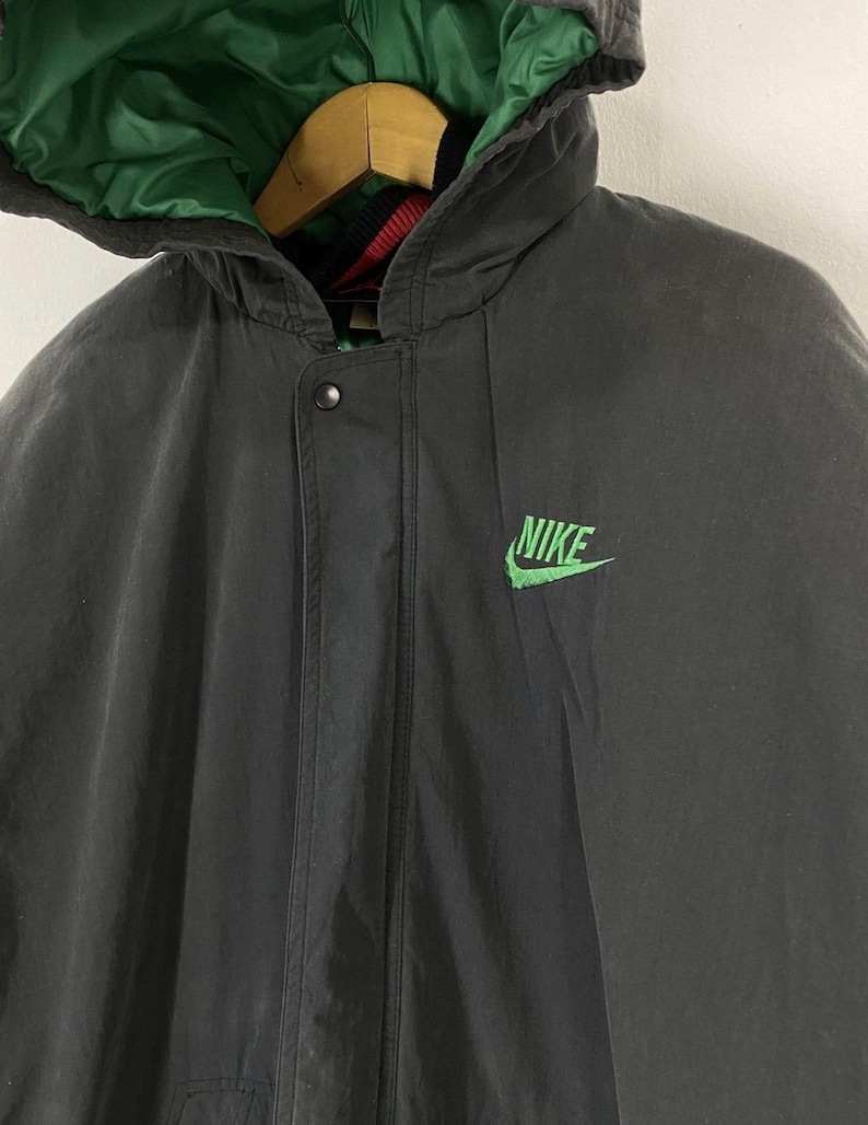 Pu&ograve; includere: Una giacca nera con cappuccio, fodera verde e un logo Nike verde sul davanti. La giacca ha una chiusura a zip e un bottone a pressione in alto.