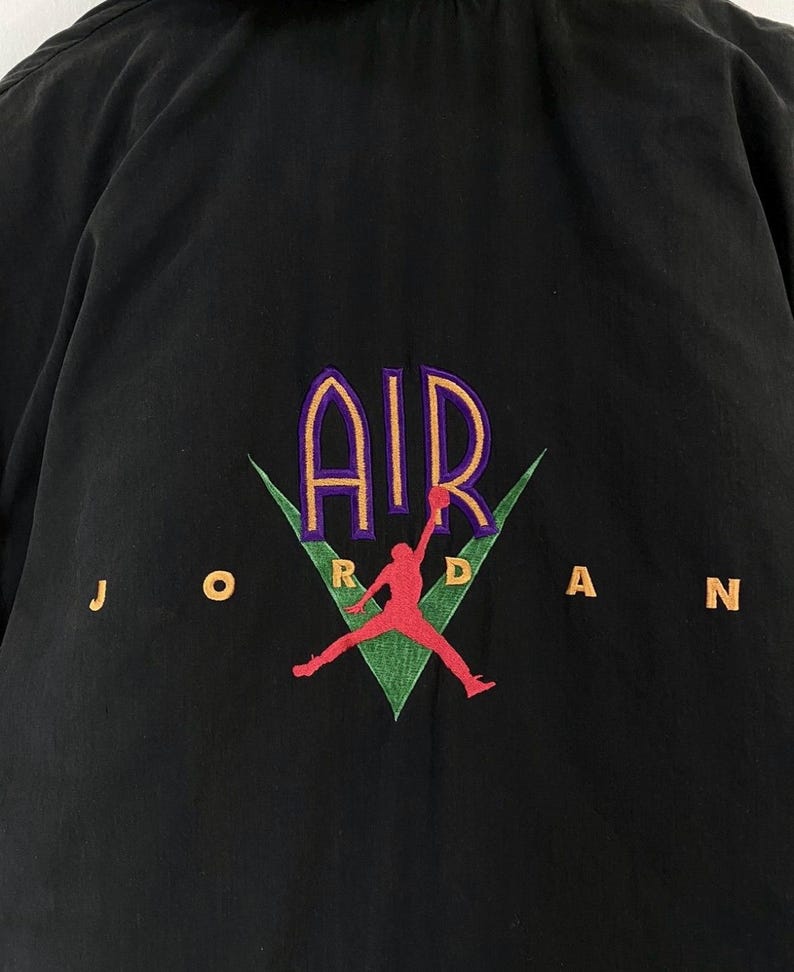 Pu&ograve; includere: Camicia nera con bottoni e un logo ricamato verde, viola e arancione che dice "AIR JORDAN" con una silhouette di un giocatore di basket.