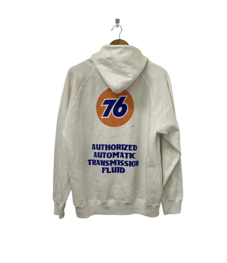 Puede incluir: Sudadera con capucha blanca con un gran c&iacute;rculo naranja que contiene el n&uacute;mero 76 en azul. El texto "AUTHORIZED AUTOMATIC TRANSMISSION FLUID" est&aacute; impreso en azul debajo del c&iacute;rculo.