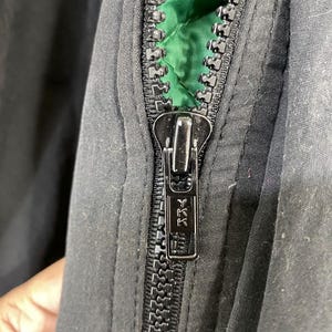 Pu&ograve; includere: Primo piano di una cerniera nera con un cursore argentato. La cerniera &egrave; parzialmente aperta, rivelando una fodera verde.