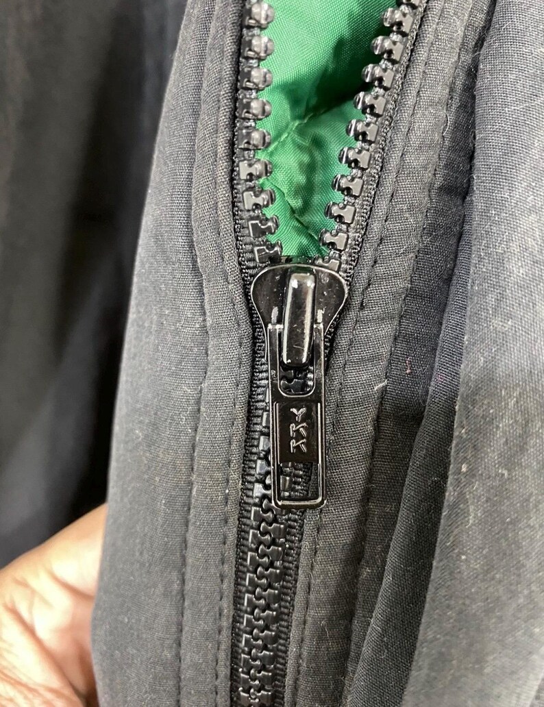 Pu&ograve; includere: Primo piano di una cerniera nera con un cursore argentato. La cerniera &egrave; parzialmente aperta, rivelando una fodera verde.